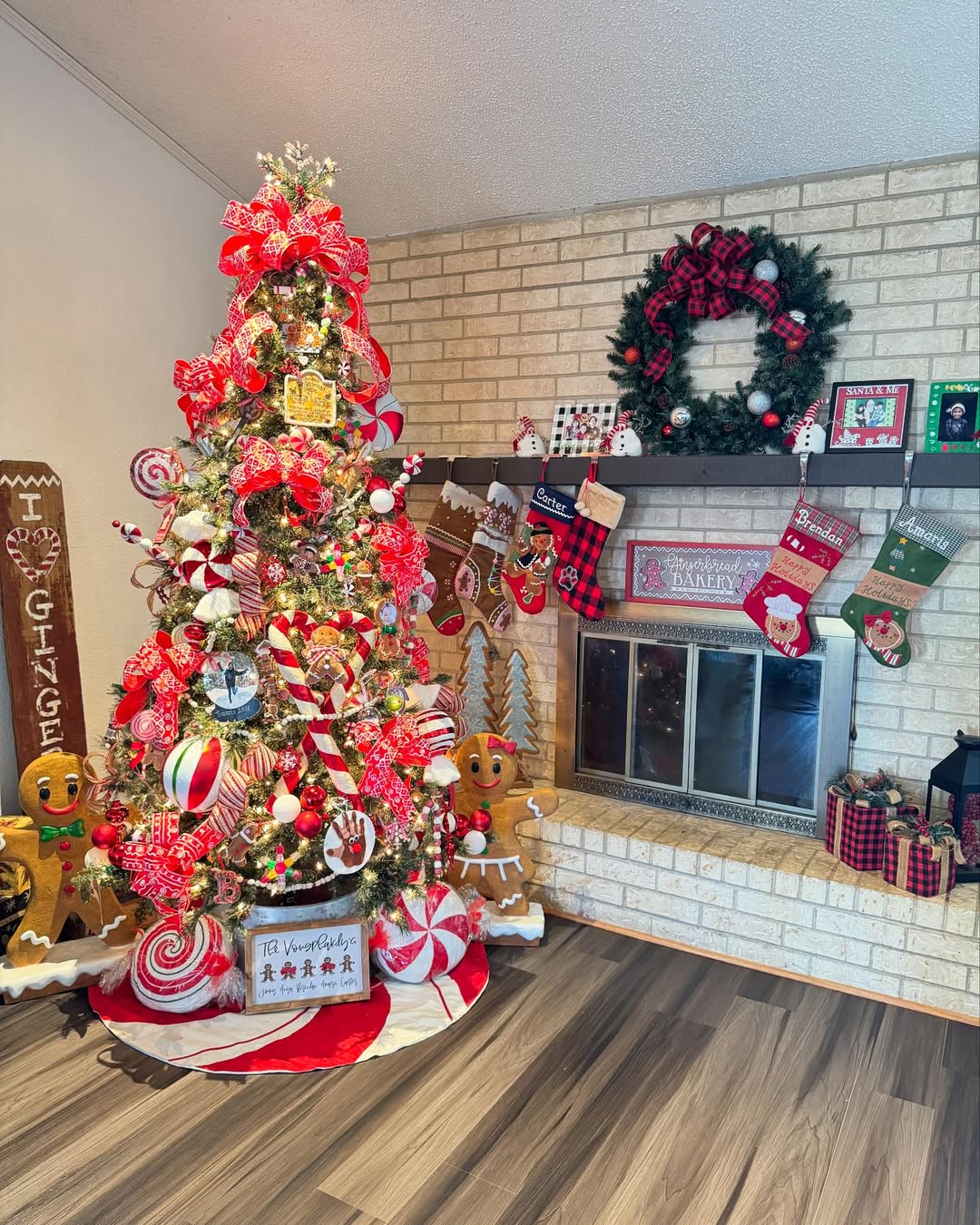 A Christmas tree decorated with red and white ornaments stands next to a fireplace with hanging stockings, a wreath, and charming Gingerbread Christmas decor on the mantel and floor—perfect for festive Christmas decor ideas.