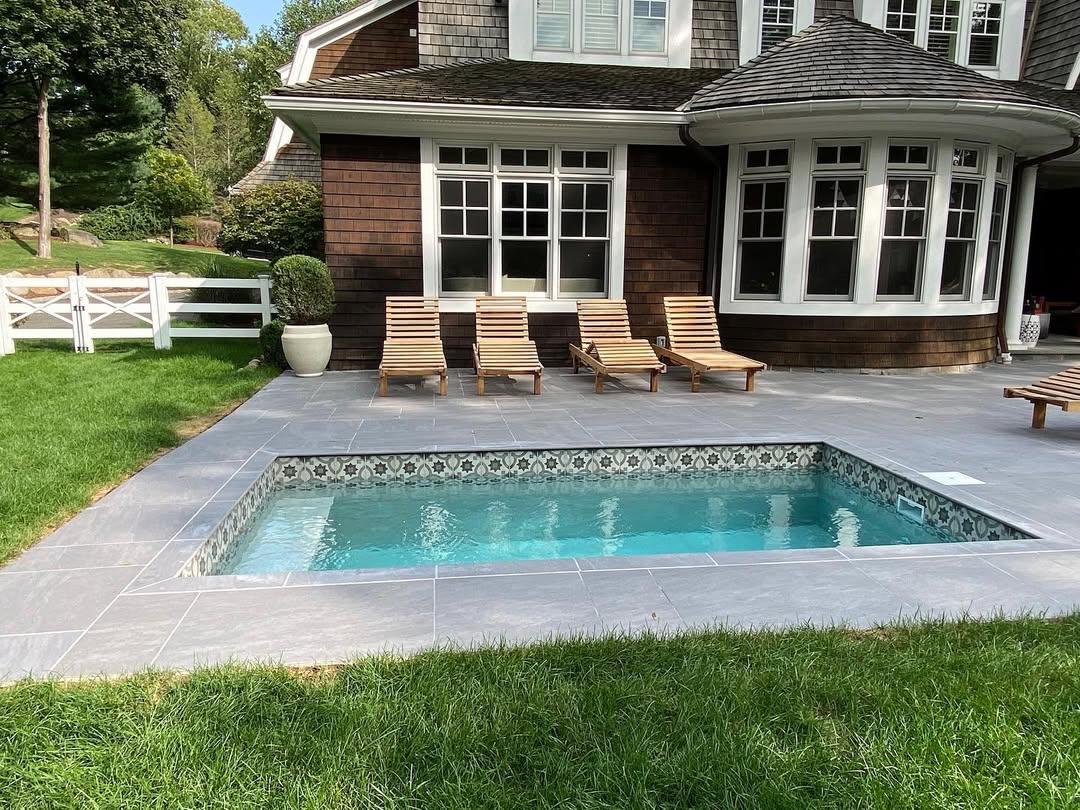 A small rectangular pool with patterned tiles is surrounded by a gray stone patio, four wooden lounge chairs, grass, and a brown house with large windows—perfect for those seeking cool pool ideas for backyard pools.