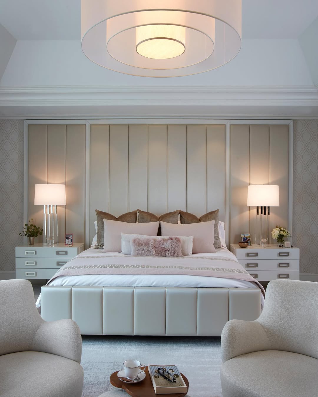 A modern bedroom features a large upholstered bed with a stylish bedroom headboard, two bedside tables with lamps, two white armchairs, and a ceiling light fixture. A tray with a teapot and cup sits on a side table.