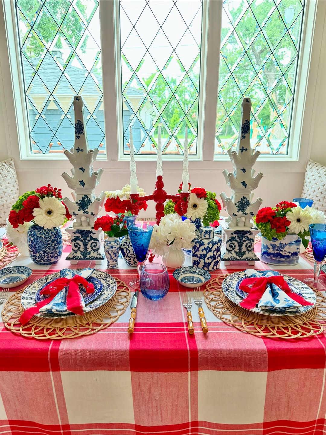 A dining table set with blue and white china, red and white flowers, blue glasses, and red napkins on a red plaid tablecloth showcases charming table decor in front of tall windows.