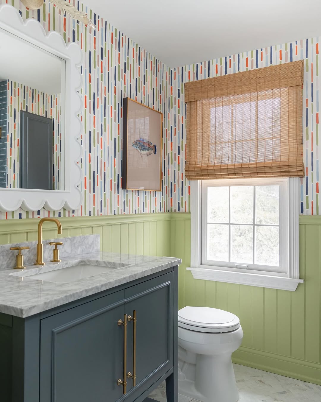 A charming kids bathroom with green wainscoting, colorful vertical striped wallpaper, a marble-topped vanity with brass fixtures, a bamboo window shade, and framed art above the toilet.