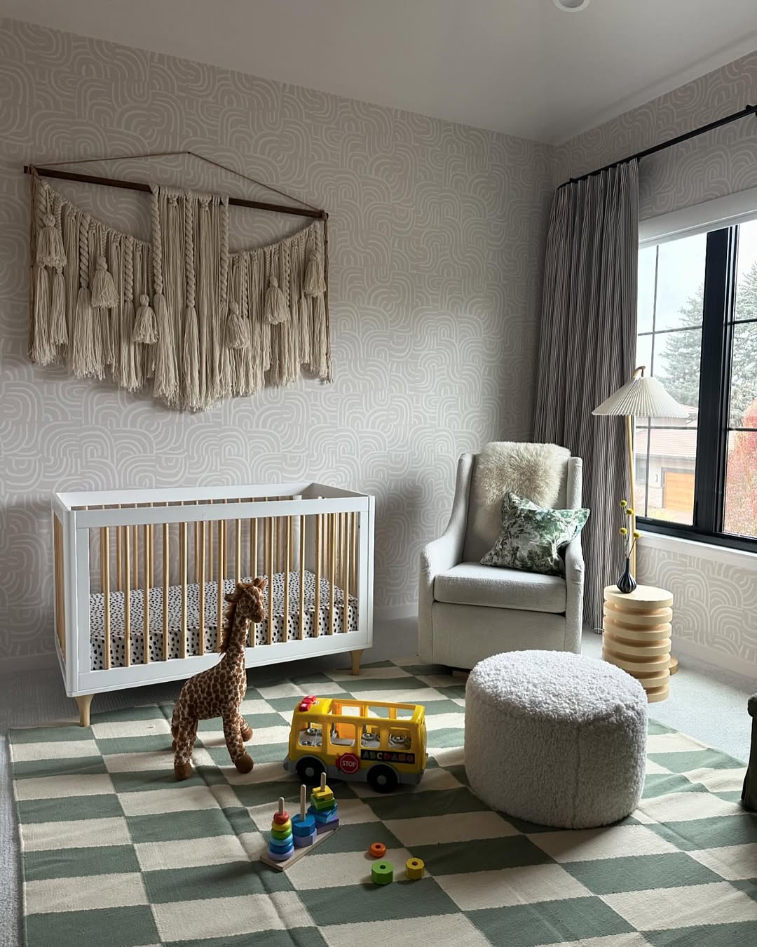 A modern nursery with a crib, stuffed giraffe, armchair, pouf, floor lamp, patterned rug, and various toys—perfect inspiration for 2025 baby boy nursery ideas and ideal for any first-time mom designing a cozy space near the window.