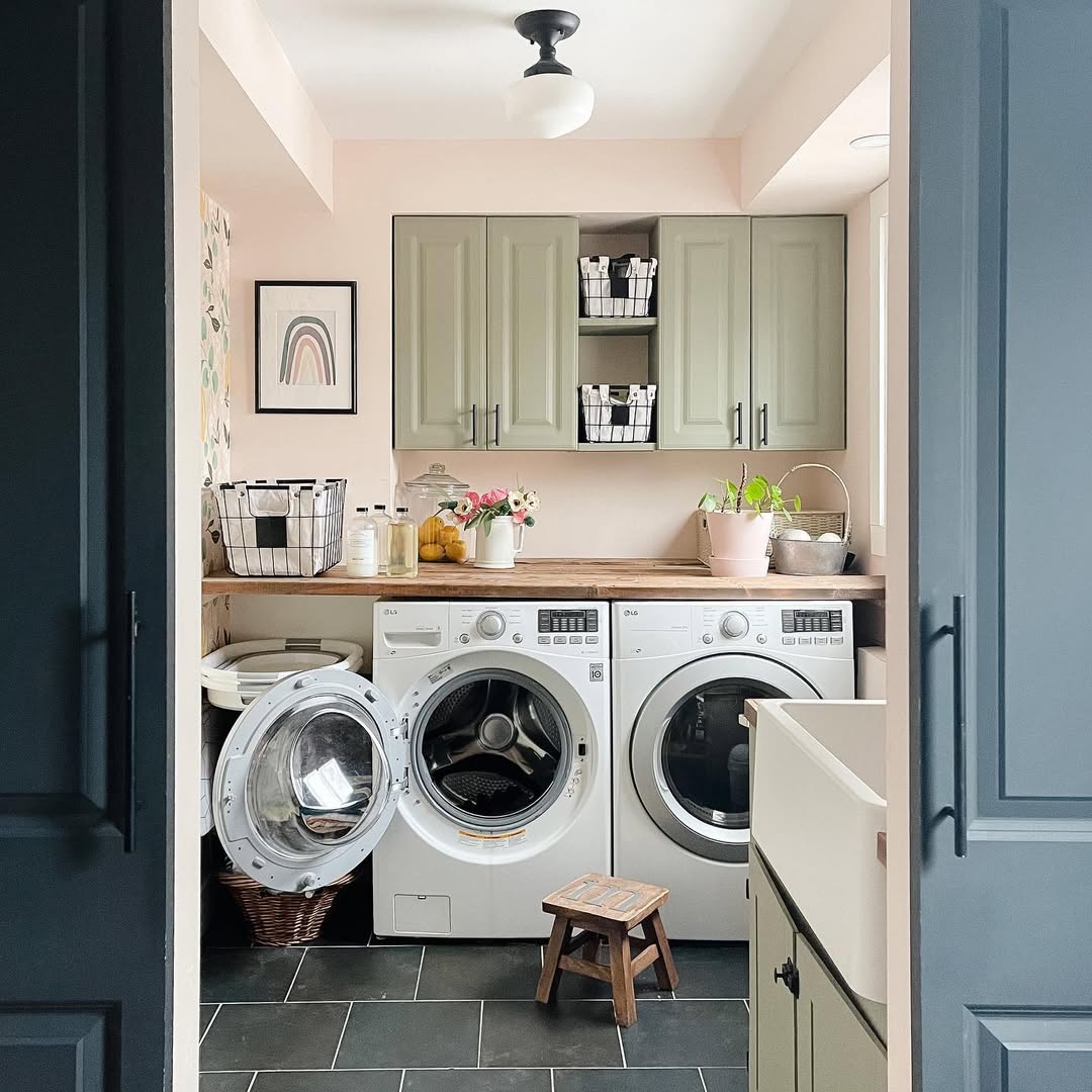 A laundry room with green laundry room cabinets, two front-loading machines (one with the door open), baskets, a wooden countertop, plants, and a small step stool.