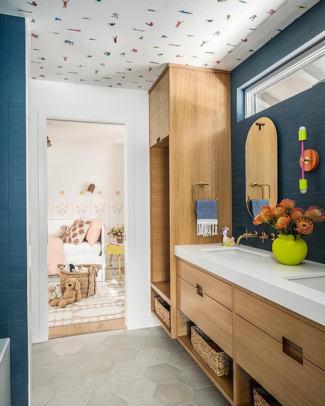 Modern kid bathroom with light wood cabinetry, dual sinks, a green vase with flowers, and a view into a cozy bedroom adorned with toys and pastel decor; whimsical patterned ceiling overhead.