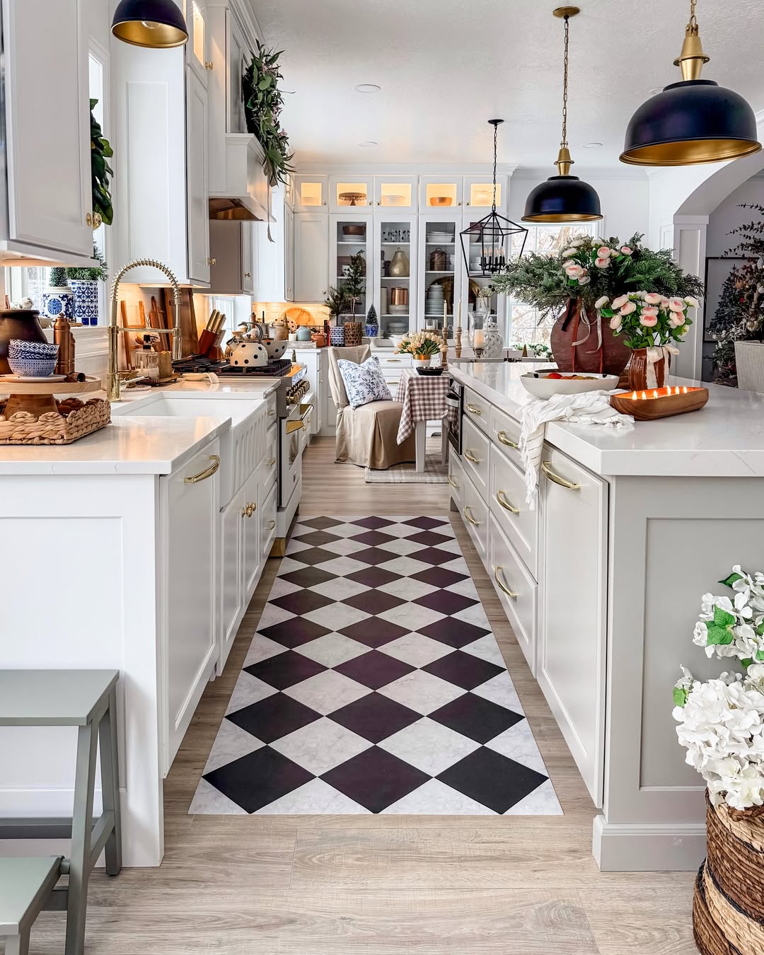 A modern farmhouse kitchen with white cabinets, gold hardware, a black-and-white checkered floor runner, pendant lights, and floral arrangements leads to a dining area—perfect for inspiring new farmhouse kitchen ideas.