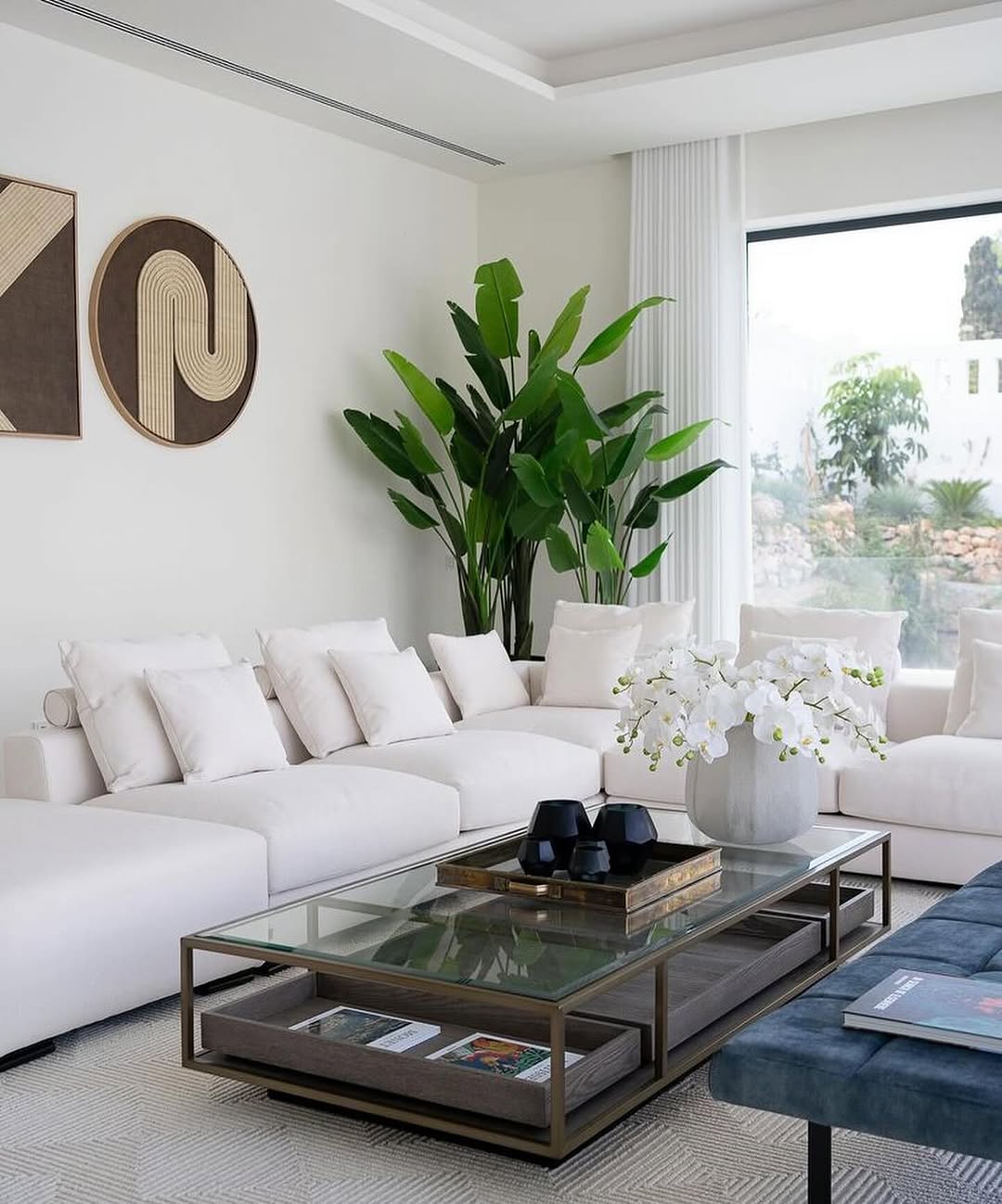 Modern organic modern living room with a white sectional sofa, large green plant, glass coffee table with decor and books, wall art, and a large window letting in natural light.