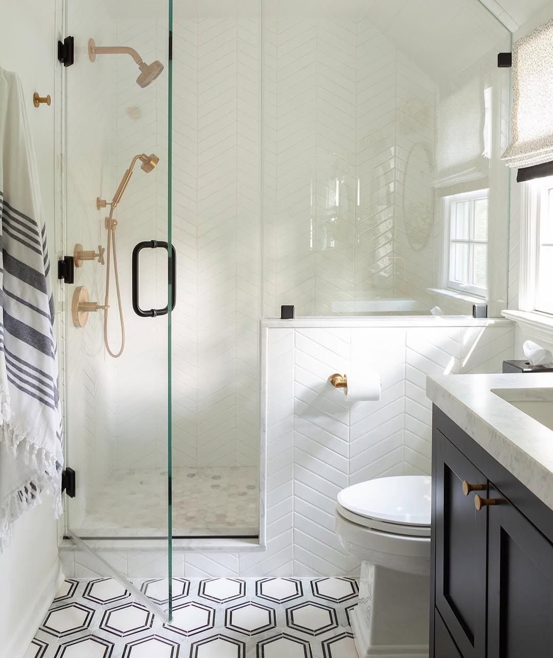 Spa-worthy bathroom featuring a glass-enclosed shower, gold fixtures, white herringbone tiles, hexagonal floor tiles, toilet, and a black vanity with a white countertop—perfect for inspiring your next bathroom design.