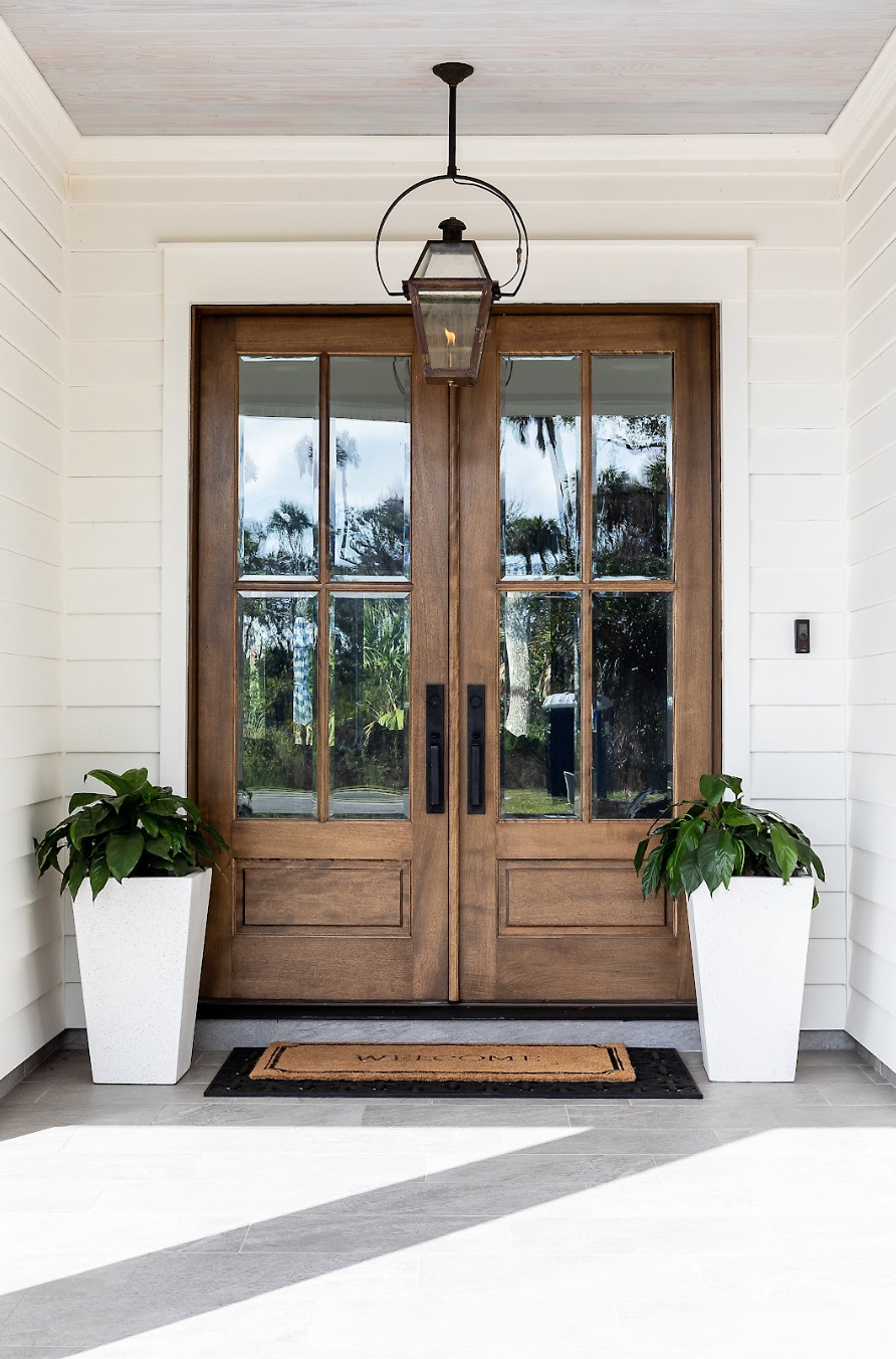 Double Door Entryway featuring wooden doors with glass panes, white siding, a welcome mat, two potted plants in white planters, and a black lantern-style light fixture above.