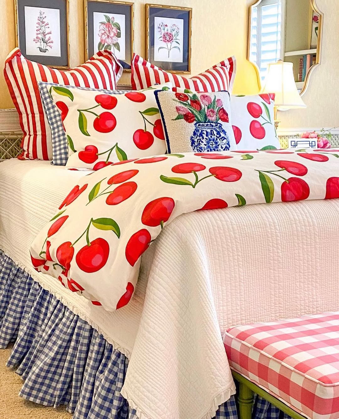 A cherry bedroom featuring white and red cherry-patterned bedding, red-and-white striped pillows, a blue gingham bed skirt, a checkered bench, and framed floral art offers fresh inspiration for charming cherry bedroom ideas.