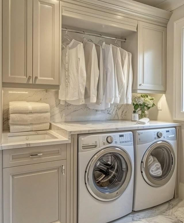 A cozy small laundry room features a washing machine and dryer, white shirts neatly hanging on a rod, stacked towels, and a charming flower arrangement adorning the marble countertop.