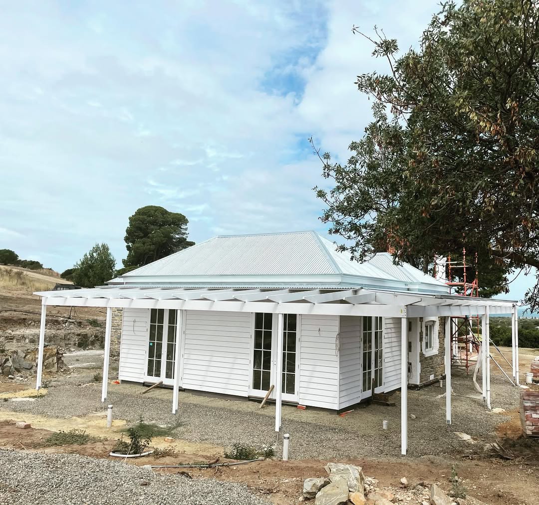 A white house with a metal roof and large covered veranda under construction showcases coastal cottage exterior inspiration for 2025, surrounded by dirt and sparse vegetation.