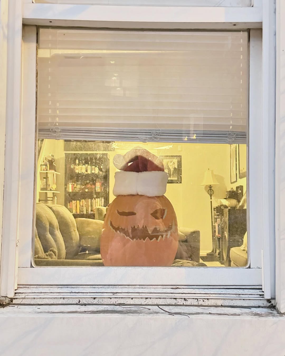 A carved pumpkin with a Santa hat sits on a couch inside a room, seen through a window with partially open blinds—offering creative Halloween window decoration ideas.