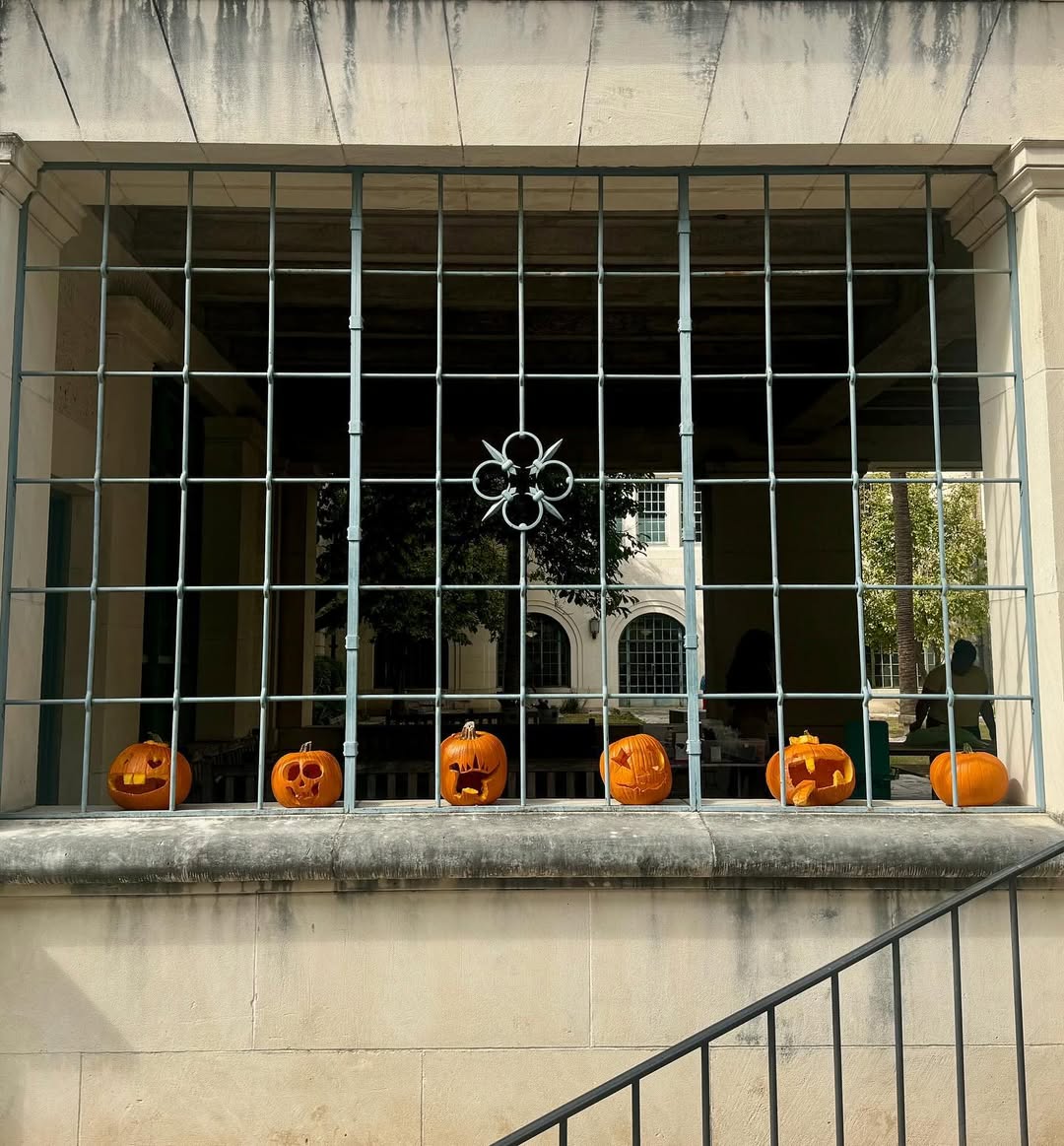 Six carved pumpkins are displayed on a stone ledge behind a metal window grille, providing creative Halloween window decoration ideas with a building and some trees visible in the background.