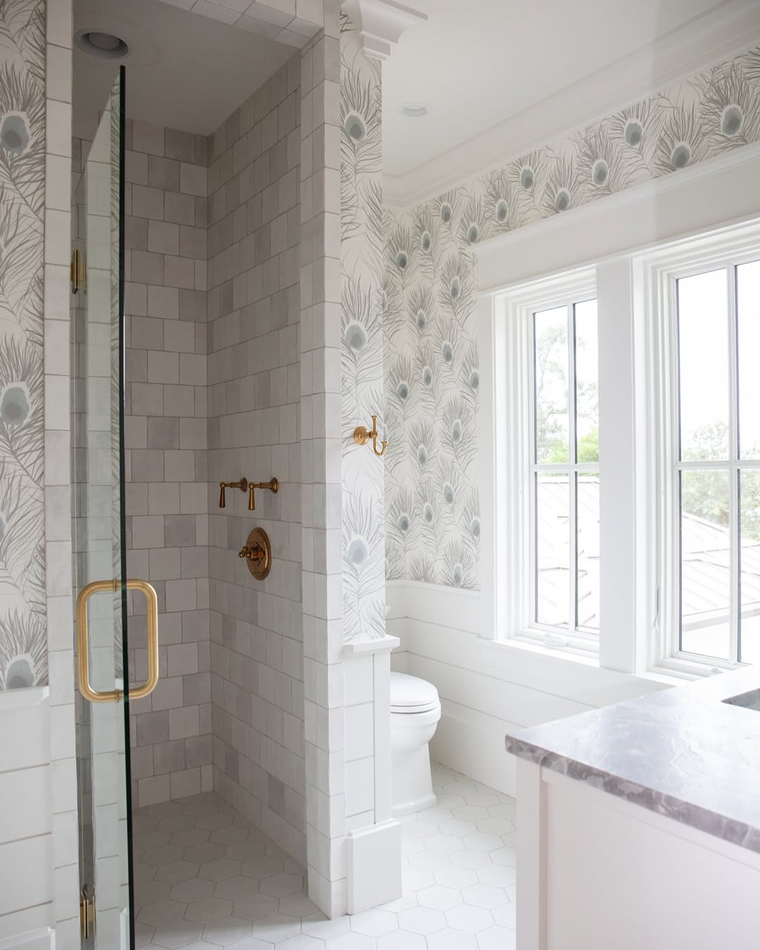 Bright bathroom design with peacock feather wallpaper, glass shower with gold fixtures, toilet by the window, and white hexagon floor tiles—perfect for spa-worthy bathroom vibes and inspiring bathroom tile ideas.