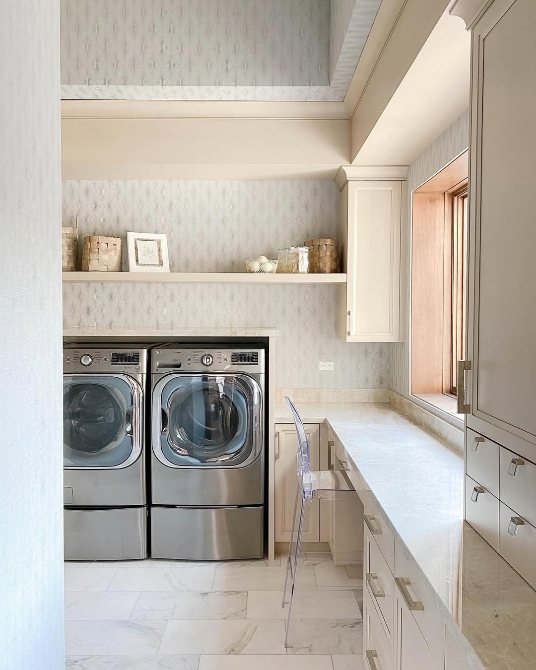 Laundry room with two front-loading machines, a clear chair at a marble counter, and inspiring laundry room cabinets, including overhead storage and floating shelf, all illuminated by a large window letting in natural light.