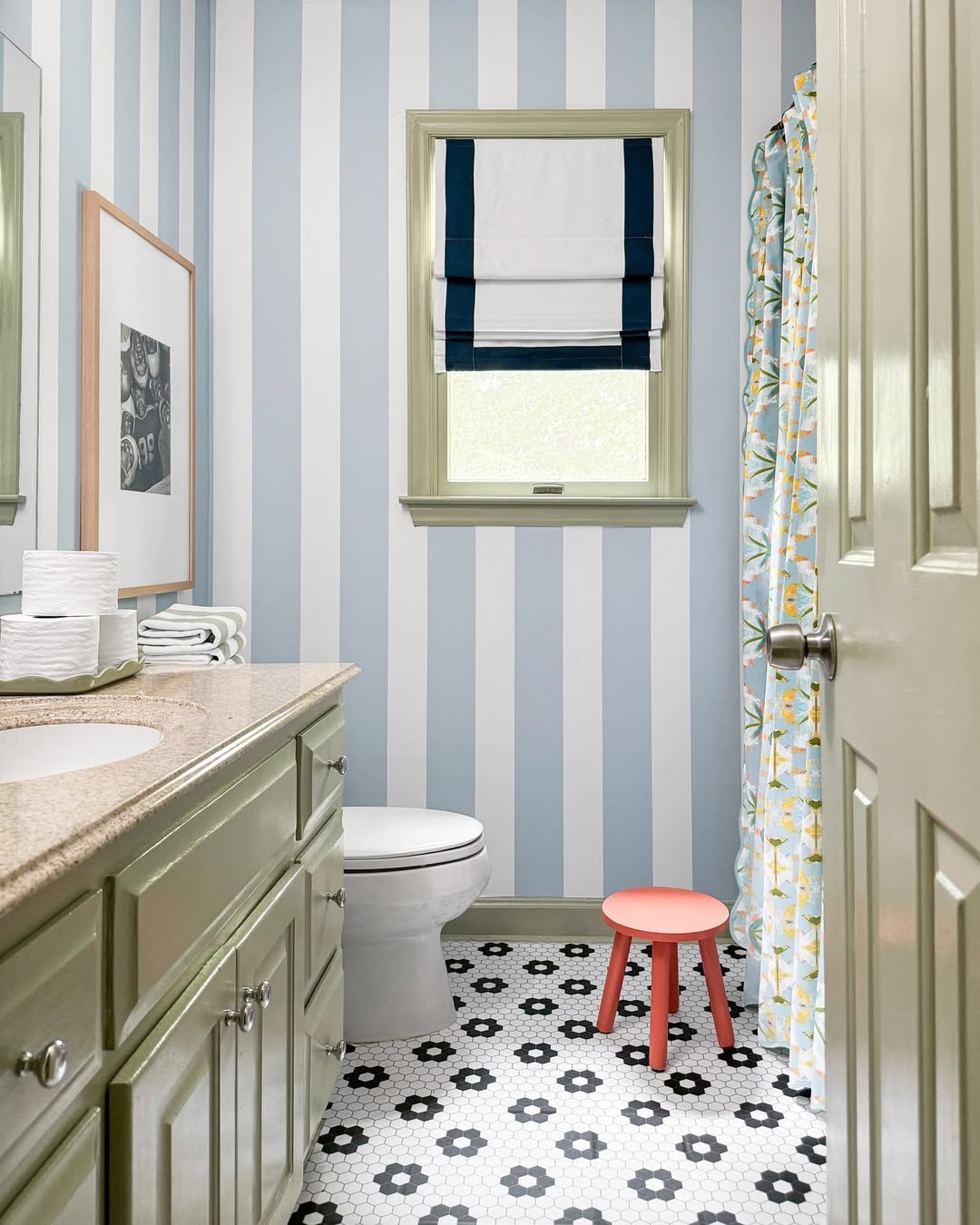 A playful kids bathroom featuring blue and white vertical striped walls, a green vanity, hexagonal black-and-white tile floor, a toilet, a red stool, and a window with a Roman shade.
