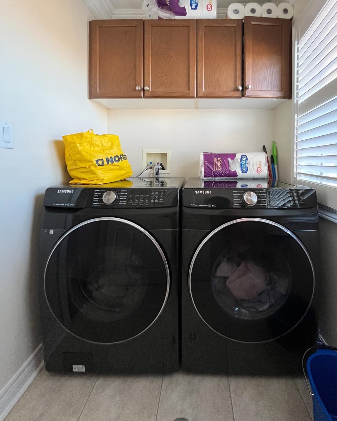 Two black Samsung front-load washer and dryer units in a laundry room with stylish Laundry Room Cabinets above, plus various household items on top, including a yellow bag and paper products.