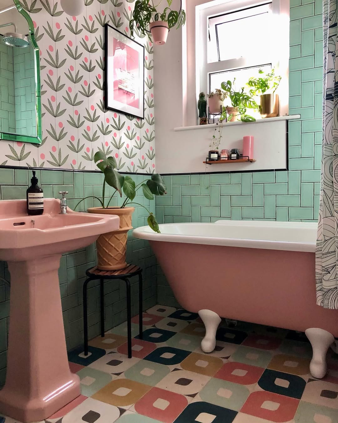 Charming pink bathroom with pedestal sink and clawfoot tub, patterned tile floor, green and pink wall tiles, leafy wallpaper, and potted plants adding a fresh touch to the windowsill and floor.