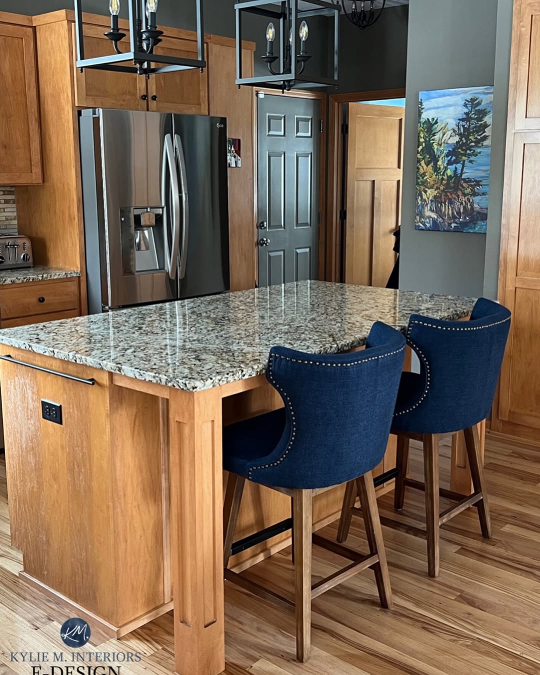 A kitchen with light wood cabinets, a granite island countertop, two blue upholstered barstools, stainless steel refrigerator, and a landscape painting on the wall showcases stunning kitchen colors that are set to lead kitchen trends.
