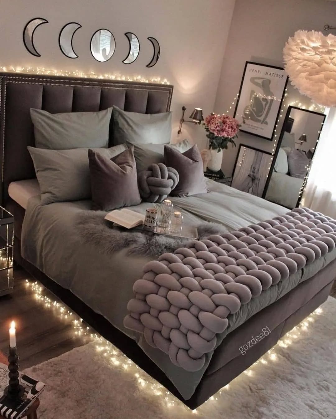 A neatly made bed with grey bedding and decorative pillows, surrounded by string lights. Showcasing inspiring women bedroom ideas, the space features moon phase wall art, flowers, candles, and chic bedroom decor touches.