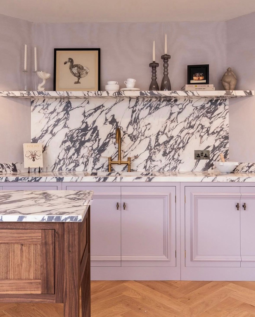 A kitchen with lilac cabinets, a marble countertop and backsplash, gold faucet, wooden island, and decorative items on an upper shelf, perfect for cottagecore decor inspiration.