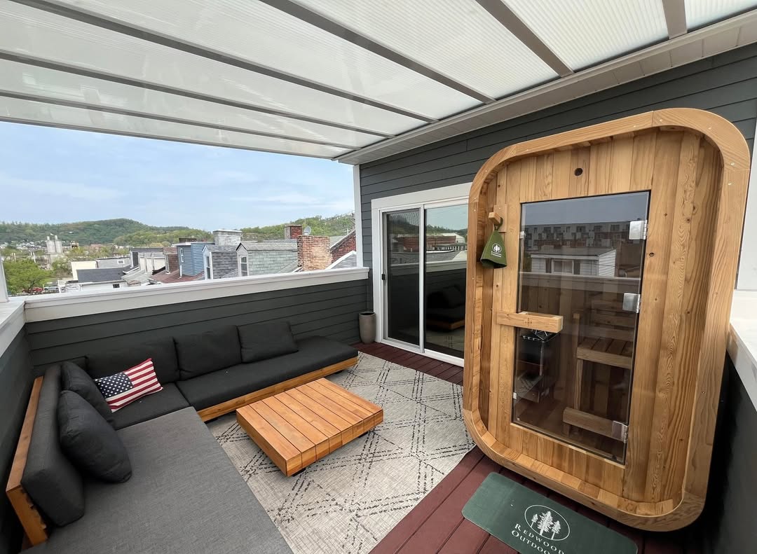 A covered rooftop patio with an outdoor sauna, gray sectional sofa, wooden coffee table, and a rug; city rooftops and hills create a scenic backdrop for those seeking gorgeous saunas and inspiring sauna ideas.