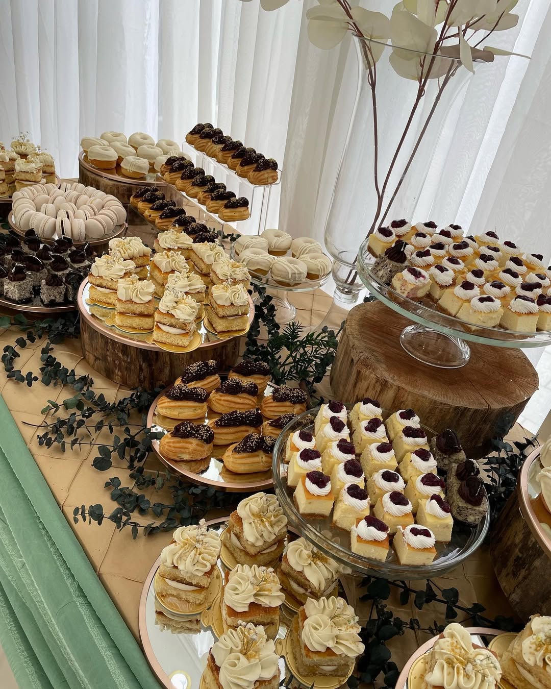 A dessert table featuring pretty desserts—assorted pastries, cakes, eclairs, and meringues—arranged on glass and wooden stands with greenery and white curtains as a charming backdrop for occasion desserts.