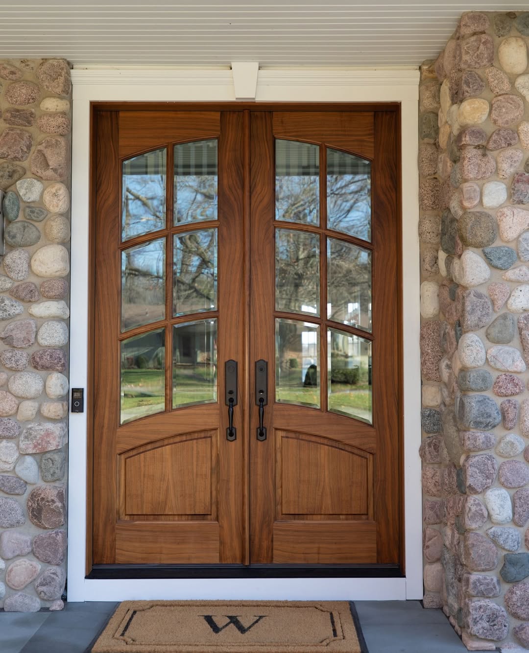 Double wooden front doors with glass panels and black handles create a stylish double door entryway, set in a stone wall. A tan doormat with the letter