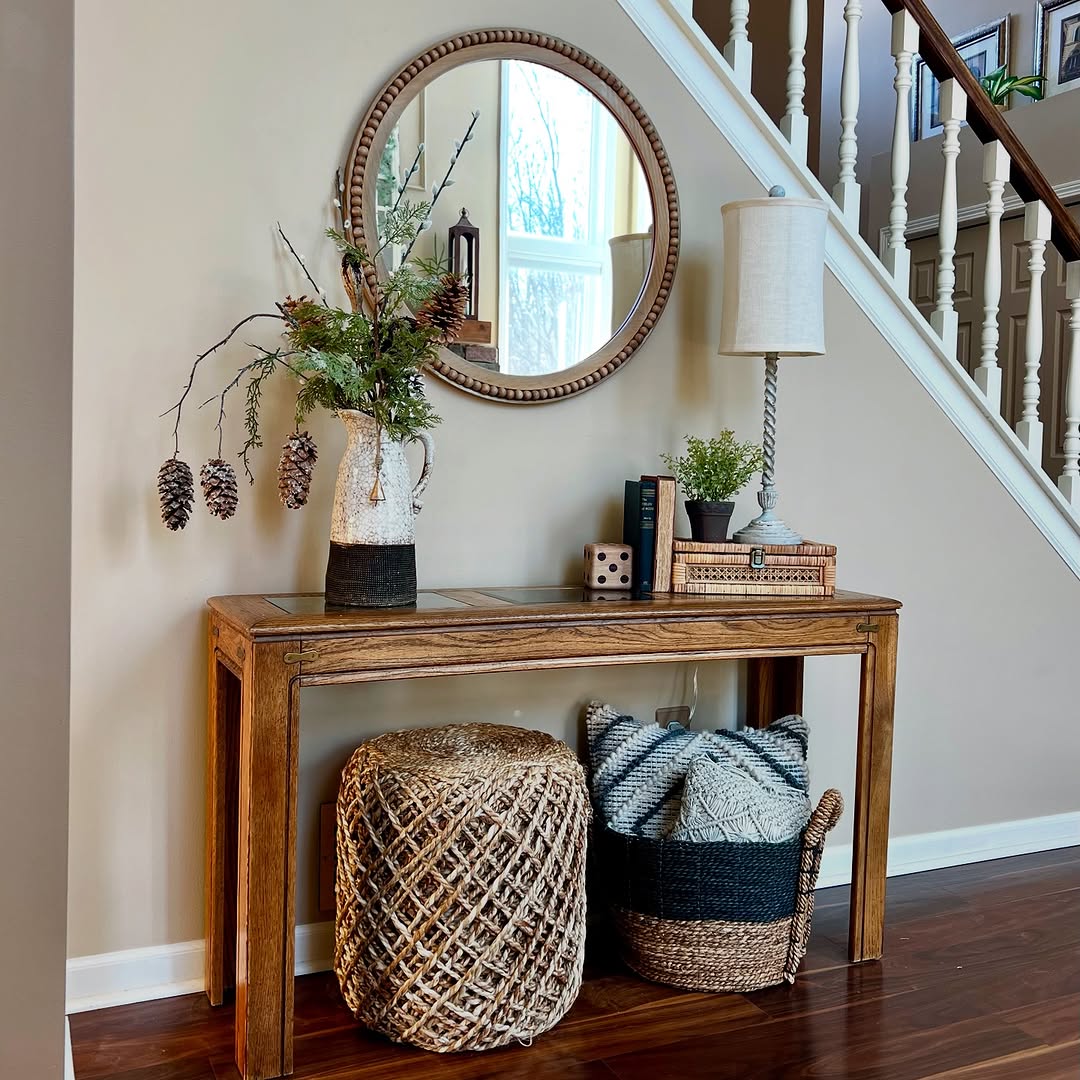 A wooden console entry table decorates the staircase wall, topped with a vase of branches, books, lamp, and decor items. Baskets sit below and a large round mirror hangs above for a welcoming touch.