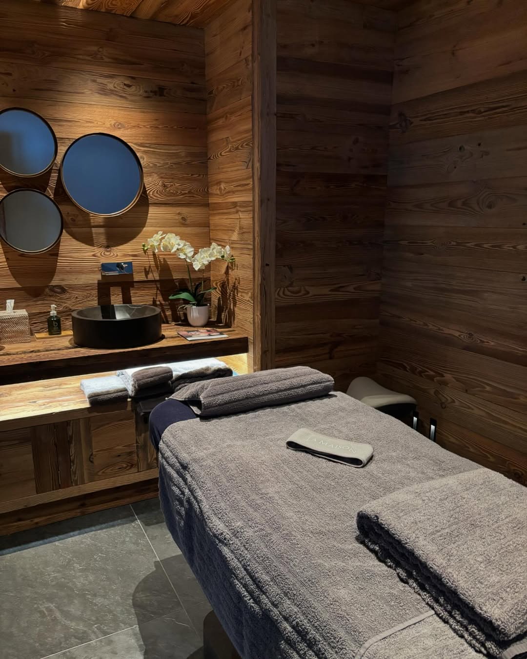 A neatly prepared massage table with folded towels in a wooden-paneled massage room, featuring a sink, mirrors, and decorative orchids on the counter offers inviting inspiration for massage room design.