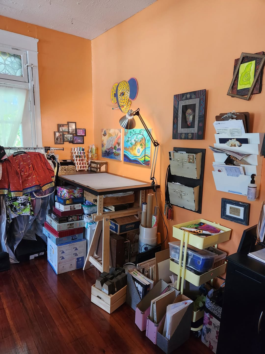 A home art studio with orange walls, art supplies, storage bins, a drafting table, wall art, and organized shelves; sunlight streams in from a window on the left—perfect inspiration for home office workshop ideas.