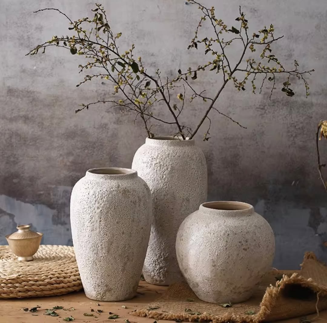 Three textured white ceramic vases on a wooden surface showcase beautiful vase decor; one vase holds thin branches with small leaves and buds. Woven mats and a small container complete this elegant arrangement.