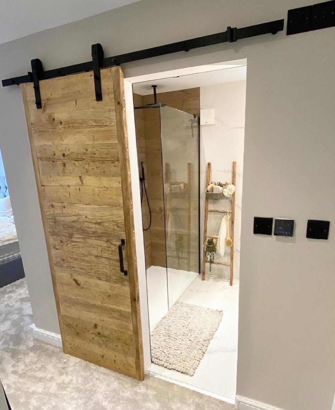 A bathroom with a glass shower and an antique bathroom door, beige carpet flooring, and a wooden ladder shelf holding towels and toiletries.