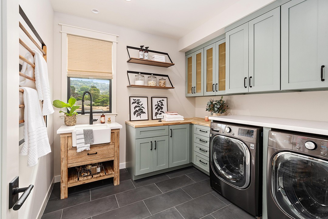 Modern laundry room with green laundry room cabinets, black washer and dryer, farmhouse sink, wooden countertops, open shelves, and decorative jars and plants. A window lets in natural light—perfect inspiration for fresh laundry room ideas.