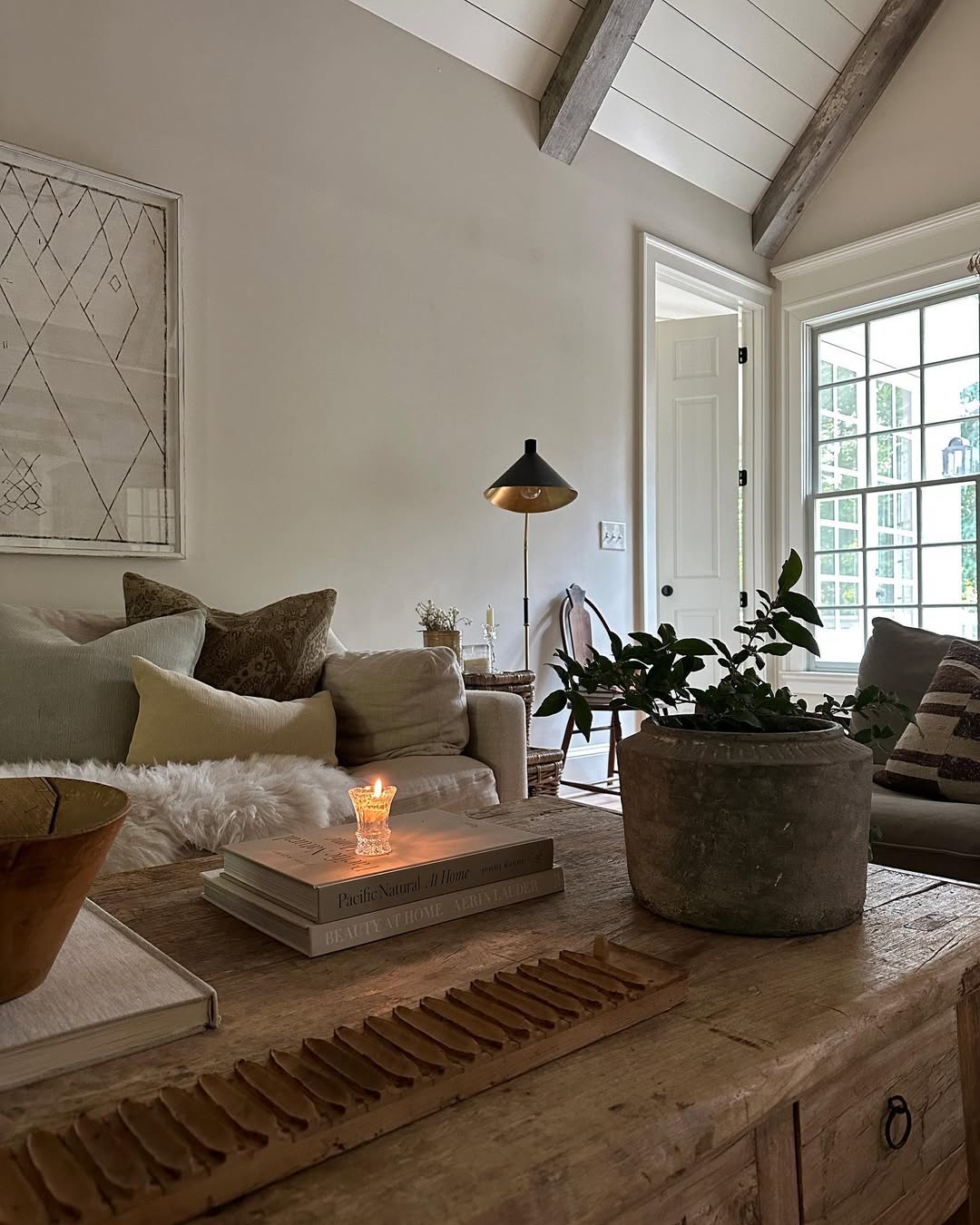 A cozy, organic modern living room with neutral tones, a sofa with pillows, a potted plant on a wooden coffee table, a lit candle, books, and a floor lamp by the window—perfect for inspiring modern living room ideas.