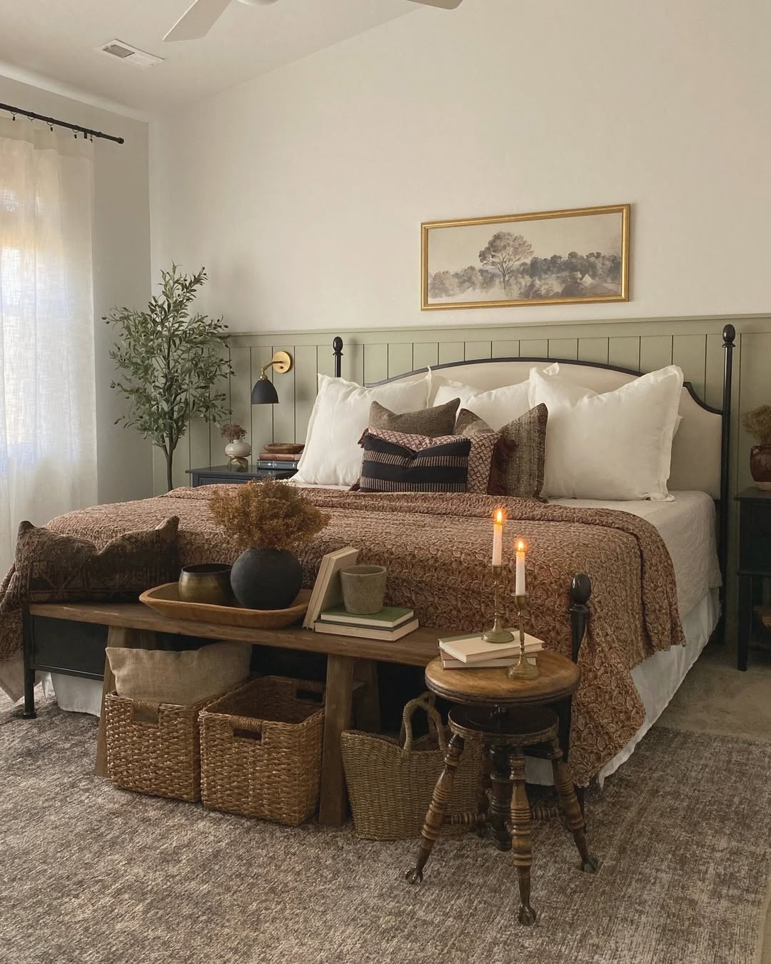 A neatly made sage green farmhouse bedroom features neutral bedding and pillows, a bench with baskets and decor, a side table with candles, and a plant in a cozy, softly lit space.