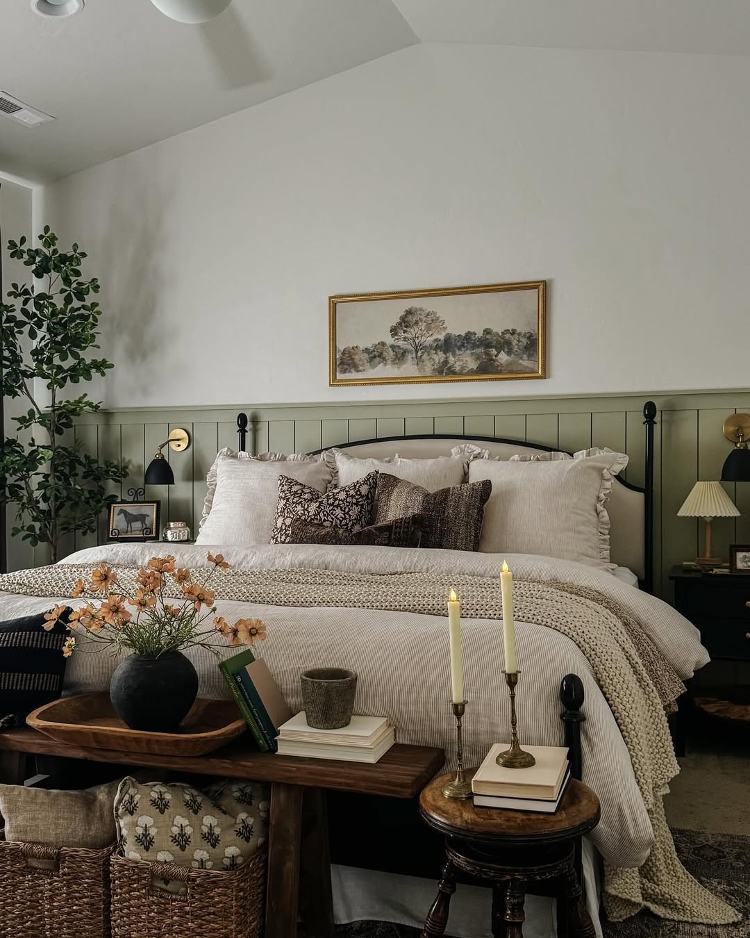 A neatly made bed with neutral linens and throw pillows is centered in a cozy sage green farmhouse bedroom, featuring wainscoting, a small bench, candles, books, and a framed landscape above the headboard.