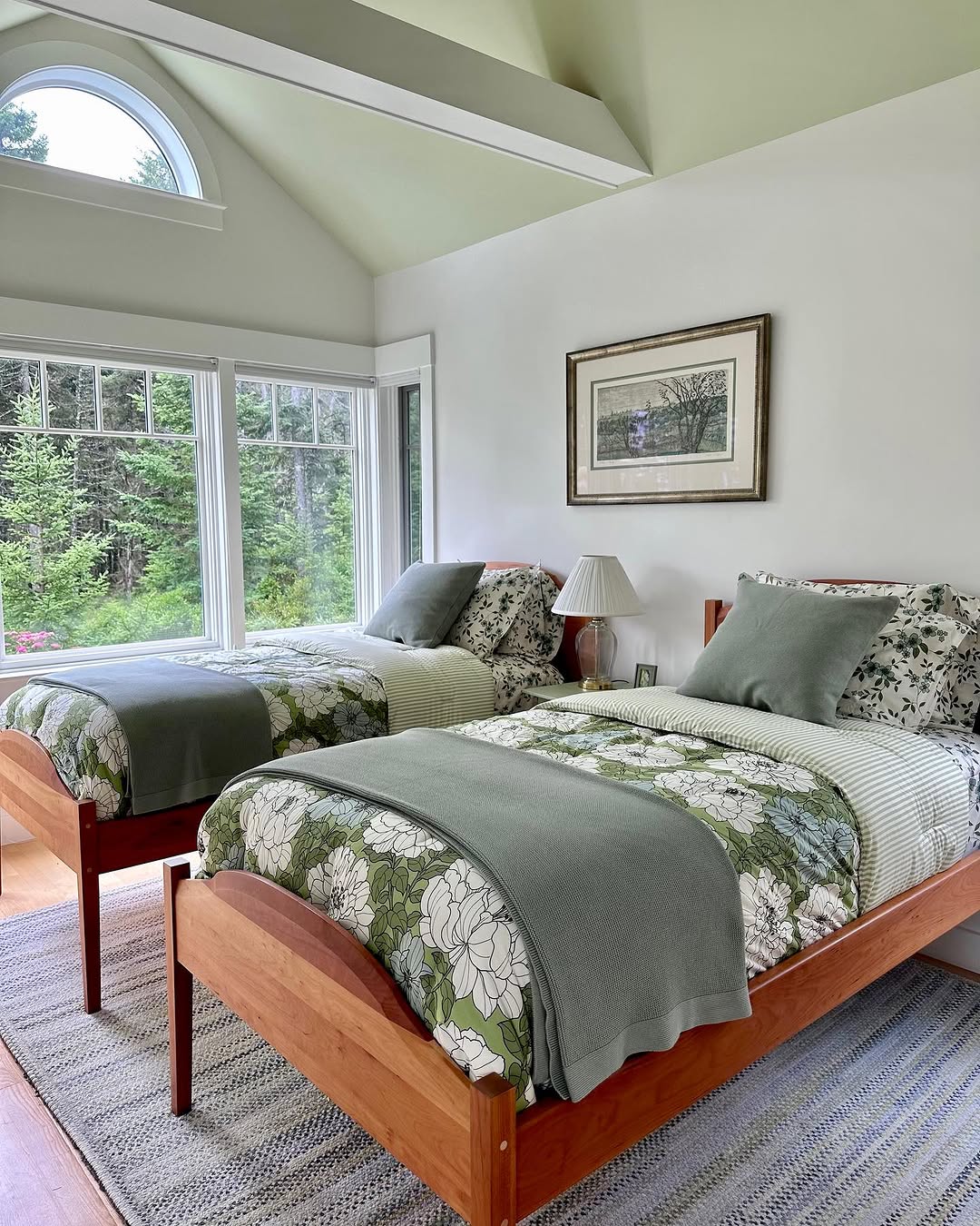 Two twin beds with sage green and floral bedding are arranged side by side in a bright farmhouse bedroom with large windows, a framed picture, and an outdoor view of trees.