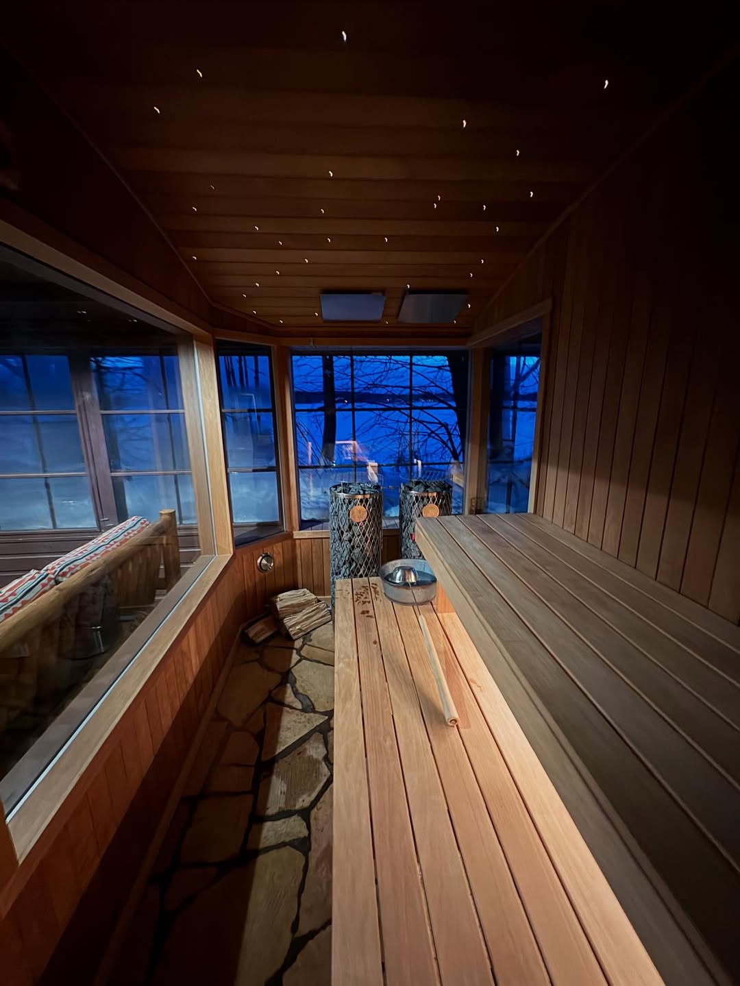 Interior of a wooden sauna with benches, a stone floor, and a heater, featuring large windows overlooking a dark blue evening scene—perfect inspiration for gorgeous saunas and outdoor wellness enthusiasts seeking new outdoor sauna ideas.
