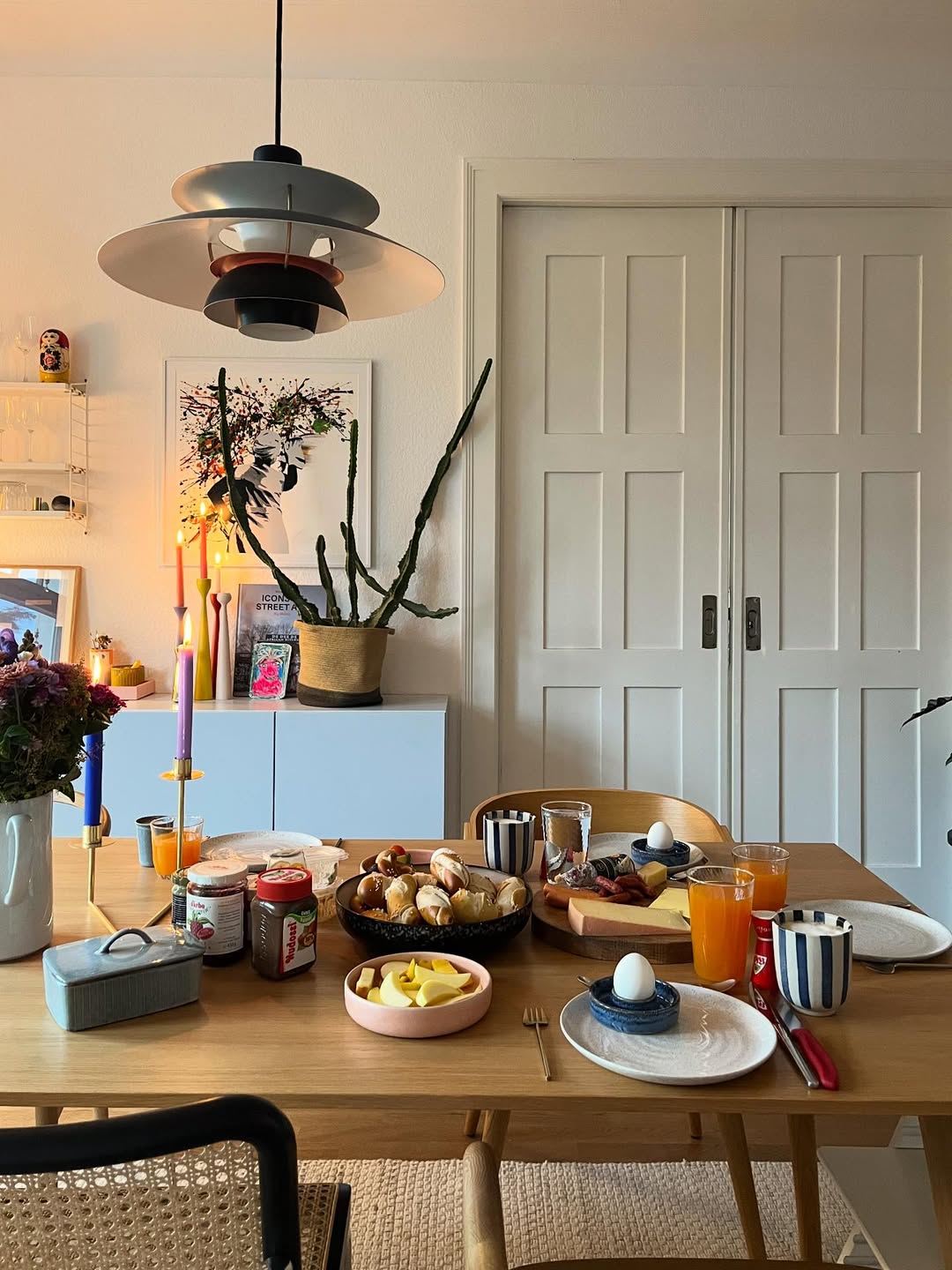 A dining table set for breakfast with eggs, bread rolls, fruit, juice, and coffee showcases stylish table settings. Candles and plants decorate the modern, well-lit room with white sliding doors in the background.
