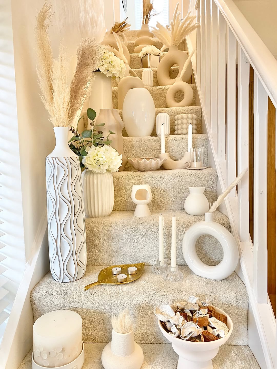 A staircase decorated with various cream and white ceramic vases, candles, dried plants, and ornamental objects arranged on each step—perfect for inspiring new vase decorating ideas.