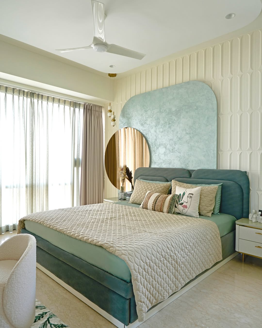 A must-see for interior lovers, this modern bedroom features a teal upholstered bed, beige quilt, decorative pillows, round mirror, and a textured accent wall—showcasing 2025’s trending bedroom color schemes.