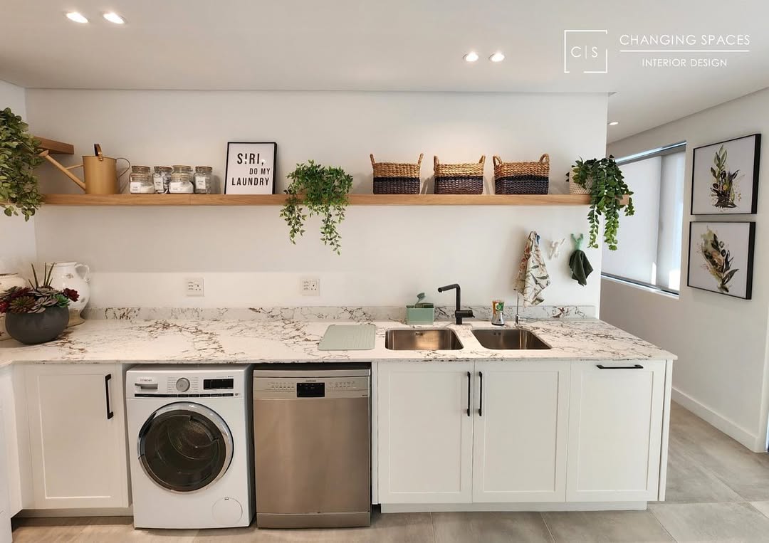 Modern boho laundry room featuring a marble countertop, white cabinets, front-loading washer, double sink, open shelves with plants, baskets, and jars, plus framed wall art—perfect for inspiring fresh boho laundry ideas.