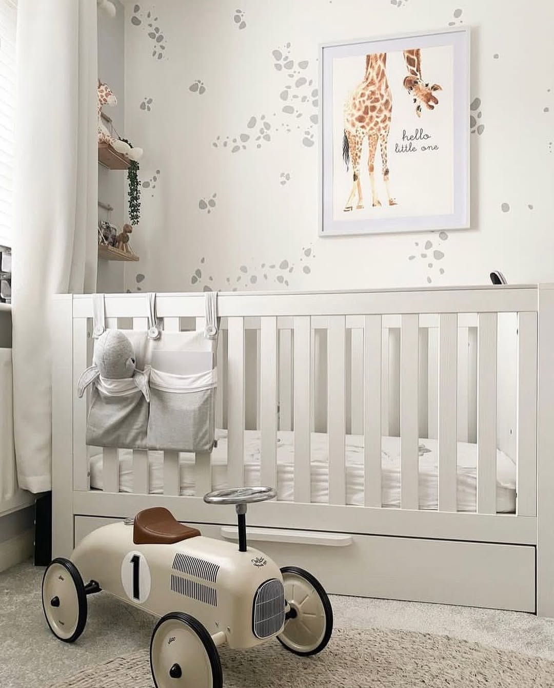A chic nursery for a first-time mom, featuring a white crib with hanging organizers, a toy car, and a framed giraffe print on a wall with gray spots in this bright baby boy nursery idea.