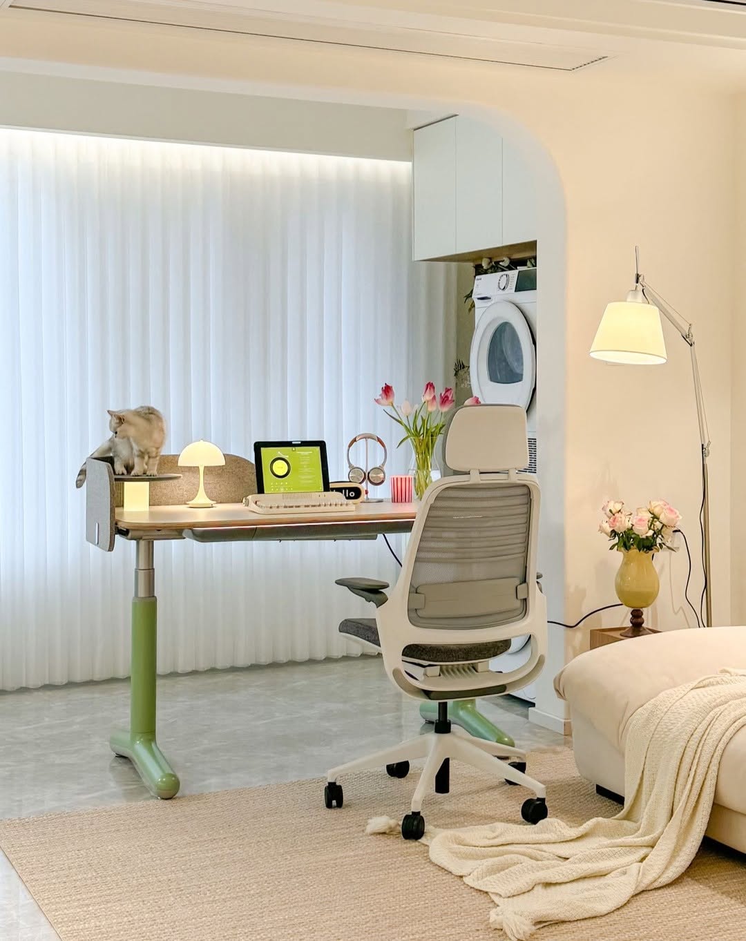 A tidy home office features a modern computer desk with a laptop, lamp, and flowers. A cat sits on the desk, while a washing machine is in the background. The space offers inspiring computer desk design ideas, with a bed and chair also present.