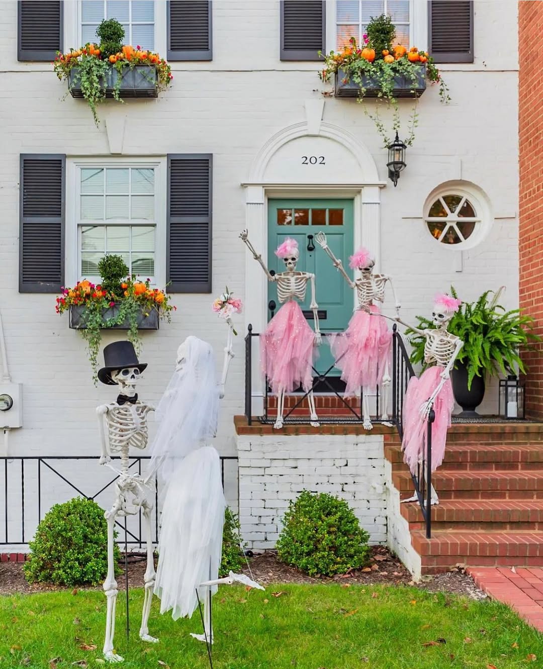 Skeleton decorations dressed as a bride, groom, and bridesmaids in pink tutus are arranged in a mock wedding scene outside a white house with flower boxes, making for unique Halloween decorations and creative Halloween window decoration ideas.