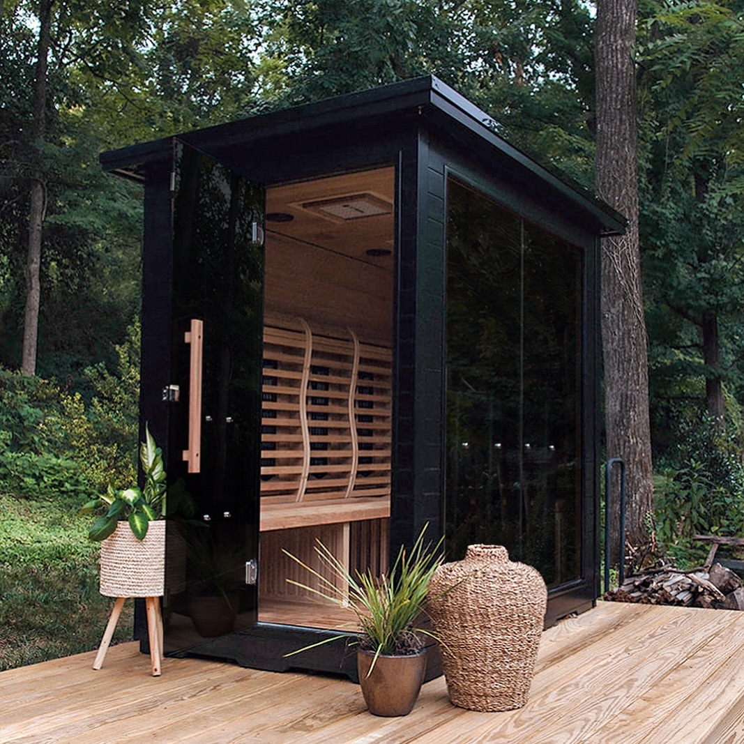 A modern black outdoor sauna with glass doors sits on a wooden deck, surrounded by potted plants and trees in a forested area—one of the most gorgeous sauna outdoor sauna ideas for nature lovers.