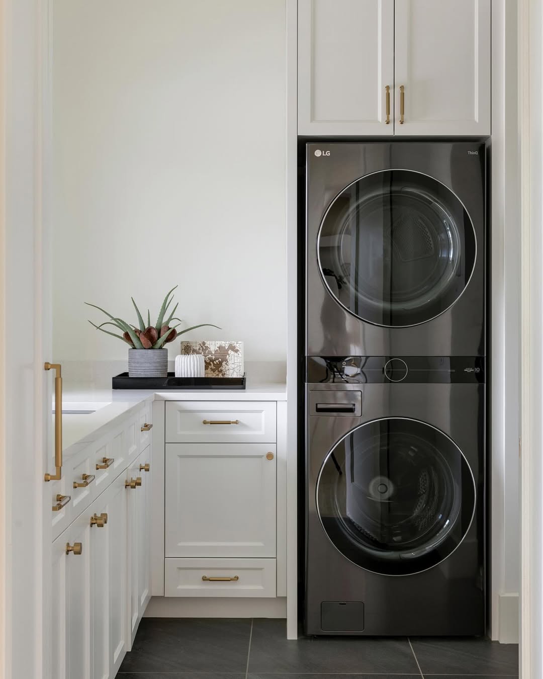 Modern laundry room with stacked black washer and dryer, stylish white laundry room cabinets with gold hardware, gray tile floor, and a countertop adorned with a plant and decor items for smart laundry storage.