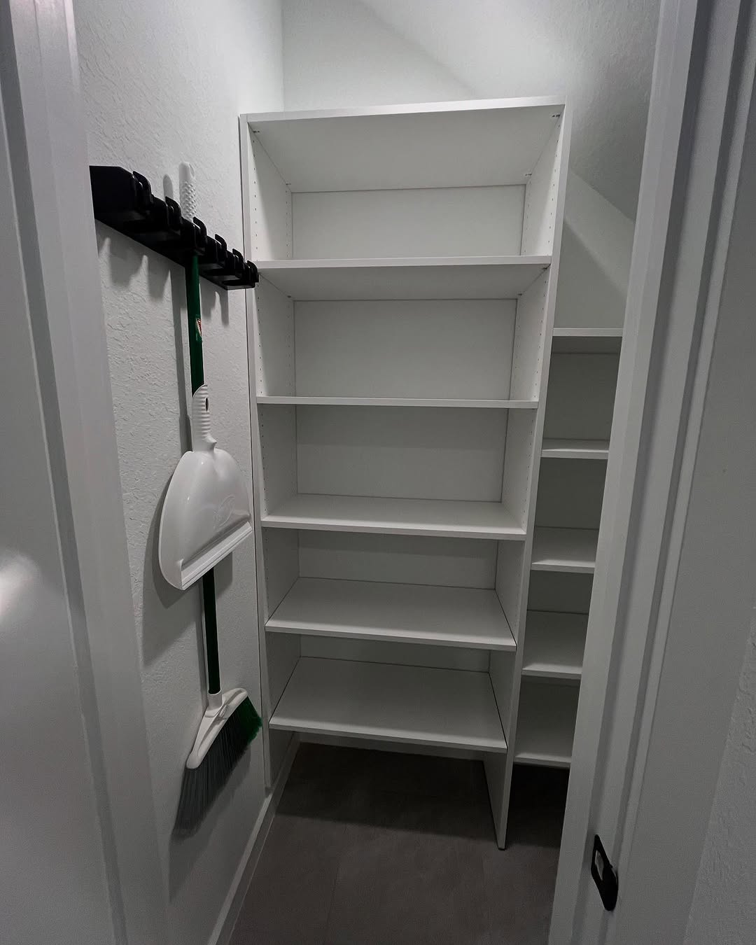 White shelving unit in a small, narrow closet with a broom and dustpan hanging on hooks—perfect for under the stairs storage or clever under stairs closet ideas.