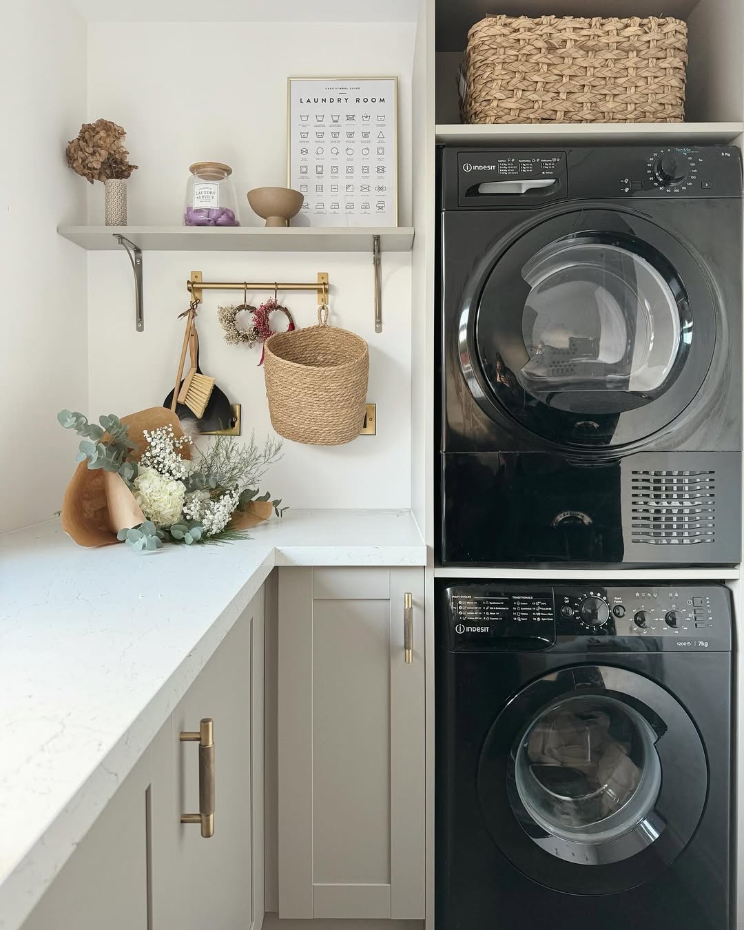 A modern stacked laundry room features a black washer and dryer, white countertops, shelves with decor, a wicker basket, and a bouquet of flowers.